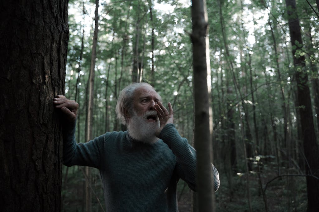 Senior man in forest shouting for assistance, wearing gray sweater, surrounded by trees.