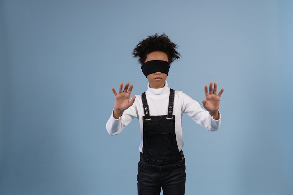 A black child in a denim jumper and white sleeves blindfolded against a blue background.